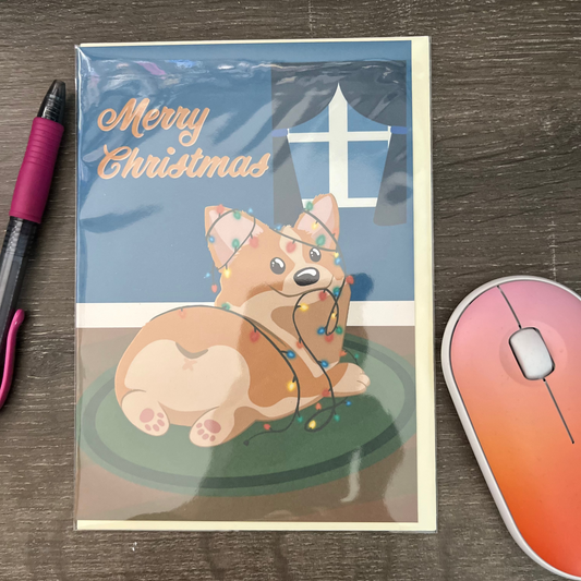Christmas card with a corgi dog surrounded by lights on a wooden surface with a pen and mouse pad.